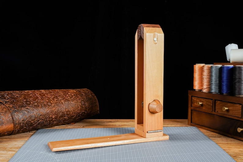 May include: A wooden leathercraft stitching pony, with a light brown finish, is displayed on a grey cutting mat. The tool has a long, vertical body and a horizontal base. Spools of thread and a leather roll are in the background.