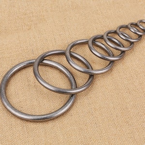 May include: A set of nine silver metal rings of varying sizes, arranged in a graduated pattern on a brown fabric background.