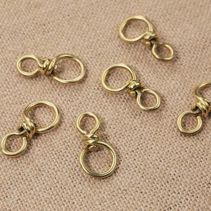 Könnte beinhalten: Sechs goldfarbene Metall-Drehverschlüsse mit einem gedrehten Knotendesign. Die Verschlüsse sind in einem Halbkreis auf einem braunen Stoffhintergrund angeordnet.