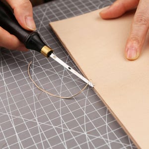 May include: A leather crafting tool with a black handle and a silver blade is used to stitch a piece of light brown leather. A curved length of thread is visible, indicating the stitching process. The background is a gray cutting mat.