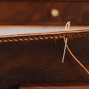May include: Close-up of a brown leather item being sewn with a needle and tan thread.