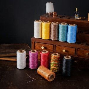 May include: Assortment of colourful spools of waxed thread for leather crafting, arranged on a wooden shelf with drawers. The spools are in various colours, including white, yellow, brown, blue, red, pink, and black.