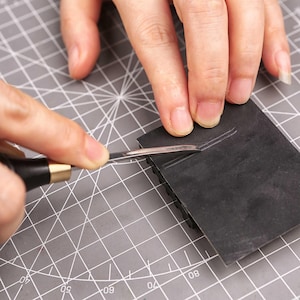 May include: A close-up shot shows a person using a craft knife to cut a dark gray piece of material. The knife has a black handle and a silver blade. The material is on a gray cutting mat with grid lines.