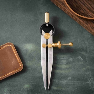 May include: A silver and brass leather crafting compass with a black knob on a green surface. The compass is used to measure and mark leather for cutting and stitching.