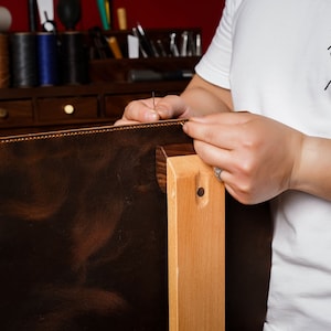 May include: A craftsman is shown sewing a dark brown leather item, possibly a bag or case, with a needle and thread. The leather is stitched to a lighter brown edge. The artisan wears a white t-shirt with a tiger graphic and the text "UNIARAI".
