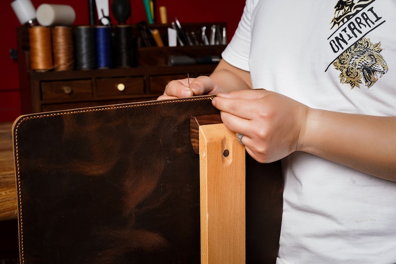 May include: A craftsman is shown sewing a dark brown leather item, possibly a bag or case, with a needle and thread. The leather is stitched to a lighter brown edge. The artisan wears a white t-shirt with a tiger graphic and the text "UNIARAI".