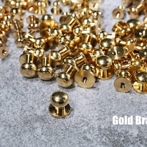 Może przedstawiać: Stosik złotych mosiężnych guzików naciskowych z okrągłą główką i gwintowaną podstawą.