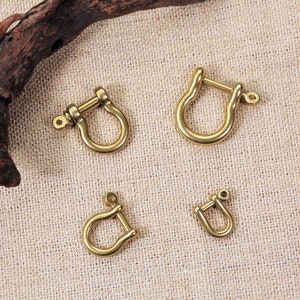 May include: Four gold-coloured metal D-rings of varying sizes. The D-rings are arranged in a grid pattern on a beige fabric surface.
