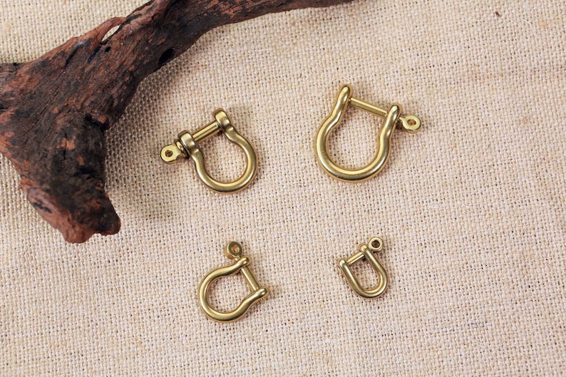 May include: Four gold-coloured metal D-rings of varying sizes. The D-rings are arranged in a grid pattern on a beige fabric surface.