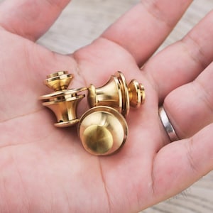 May include: Three gold-colored metal button snaps, each with a different design, are shown in a person's hand.