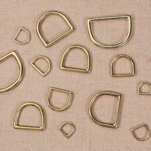 May include: Assortment of gold-toned metal D-rings of various sizes. The rings are arranged on a beige fabric background.