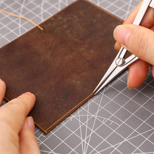 May include: A close-up of a brown leather piece being worked on with a silver metal compass. The compass is used to measure and mark the leather. The leather is on a gray cutting mat.