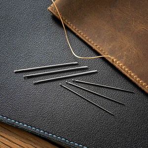 May include: A close-up of a dark blue leather surface with a light blue stitching detail. Several silver needles are scattered on the surface. A piece of brown leather with stitching detail is partially visible in the top right corner of the image.