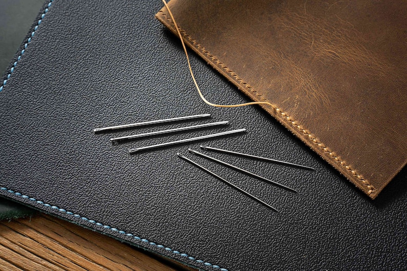 May include: A close-up of a dark blue leather surface with a light blue stitching detail. Several silver needles are scattered on the surface. A piece of brown leather with stitching detail is partially visible in the top right corner of the image.