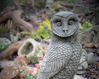 May include: A gray concrete owl statue perched on a weathered wooden stump. The owl has large, round eyes and detailed feathers.