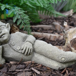 Puede incluir: Una estatua de gnomo de jardín de hormigón gris que representa a un duende dormido con un gorro puntiagudo, tumbado de lado con la cabeza apoyada en la mano. El gnomo lleva un jersey con botones y está rodeado de mantillo marrón.