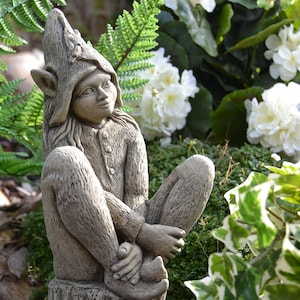 May include: A stone garden ornament depicting a seated elf wearing a pointed hat and a buttoned tunic. The elf sits on a tree stump, with crossed legs and clasped hands. The statue is a neutral grey colour, surrounded by greenery and white flowers.