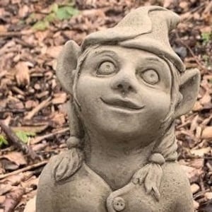 May include: A grey stone gnome figurine with a pointed hat, sitting on a log. The gnome has a friendly face and is holding its hands together in front of it.