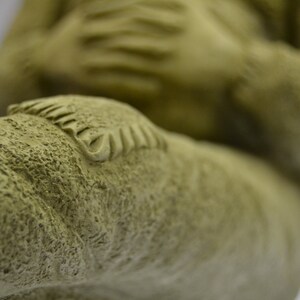 Puede incluir: Primer plano de una escultura de cer&aacute;mica verde texturizada de una persona sentada con las piernas cruzadas. La escultura tiene un dise&ntilde;o detallado en la rodilla de una pierna.