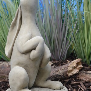 Pu&ograve; includere: Una statua di coniglio in cemento grigio con le zampe incrociate e la testa inclinata verso l'alto, in piedi su una base rotonda. La statua &egrave; circondata da fogliame verde e marrone.