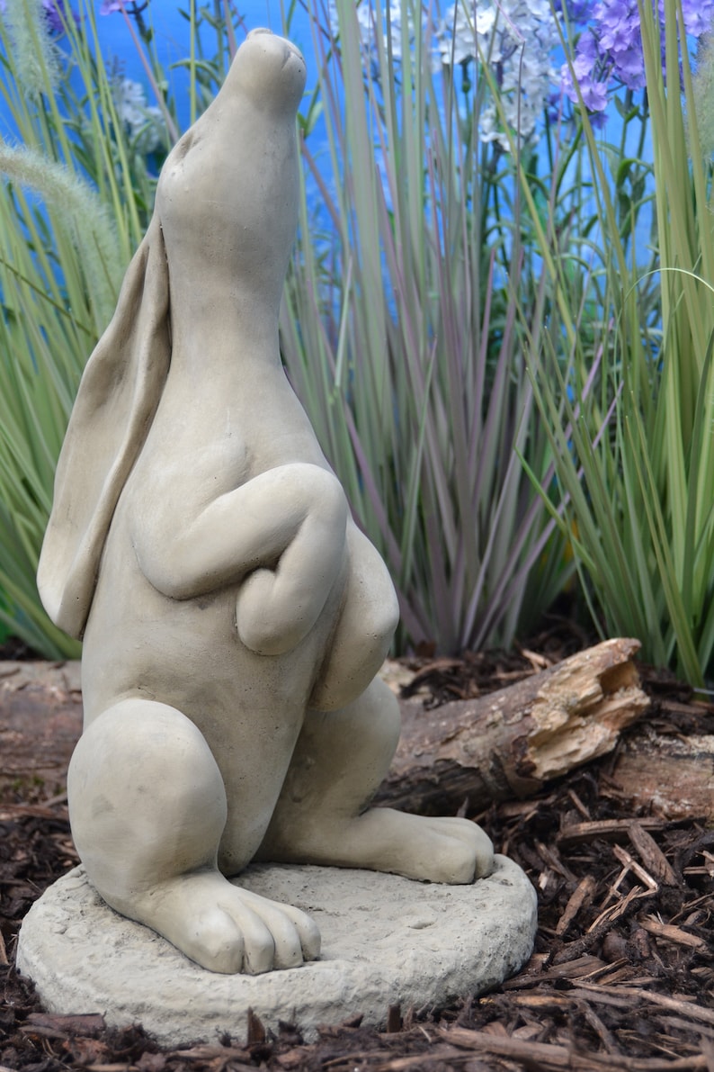 Pu&ograve; includere: Una statua di coniglio in cemento grigio con le zampe incrociate e la testa inclinata verso l'alto, in piedi su una base rotonda. La statua &egrave; circondata da fogliame verde e marrone.