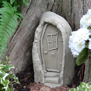 Könnte beinhalten: Eine kleine, graue, Betontür für Feen mit einer Holztür und einem Fenster. Die Tür ist in einen Baumstamm eingebaut und hat eine kleine Treppe, die zu ihr führt.