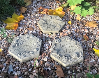 Puede incluir: Tres piedras decorativas de jardín con lemas caprichosos. La primera piedra dice "This Way for Tea" y está decorada con tazas de té y teteras. La segunda piedra dice "That Way" y está decorada con pájaros y flores. La tercera piedra dice "Don't Be Late" y está decorada con relojes y cadenas.