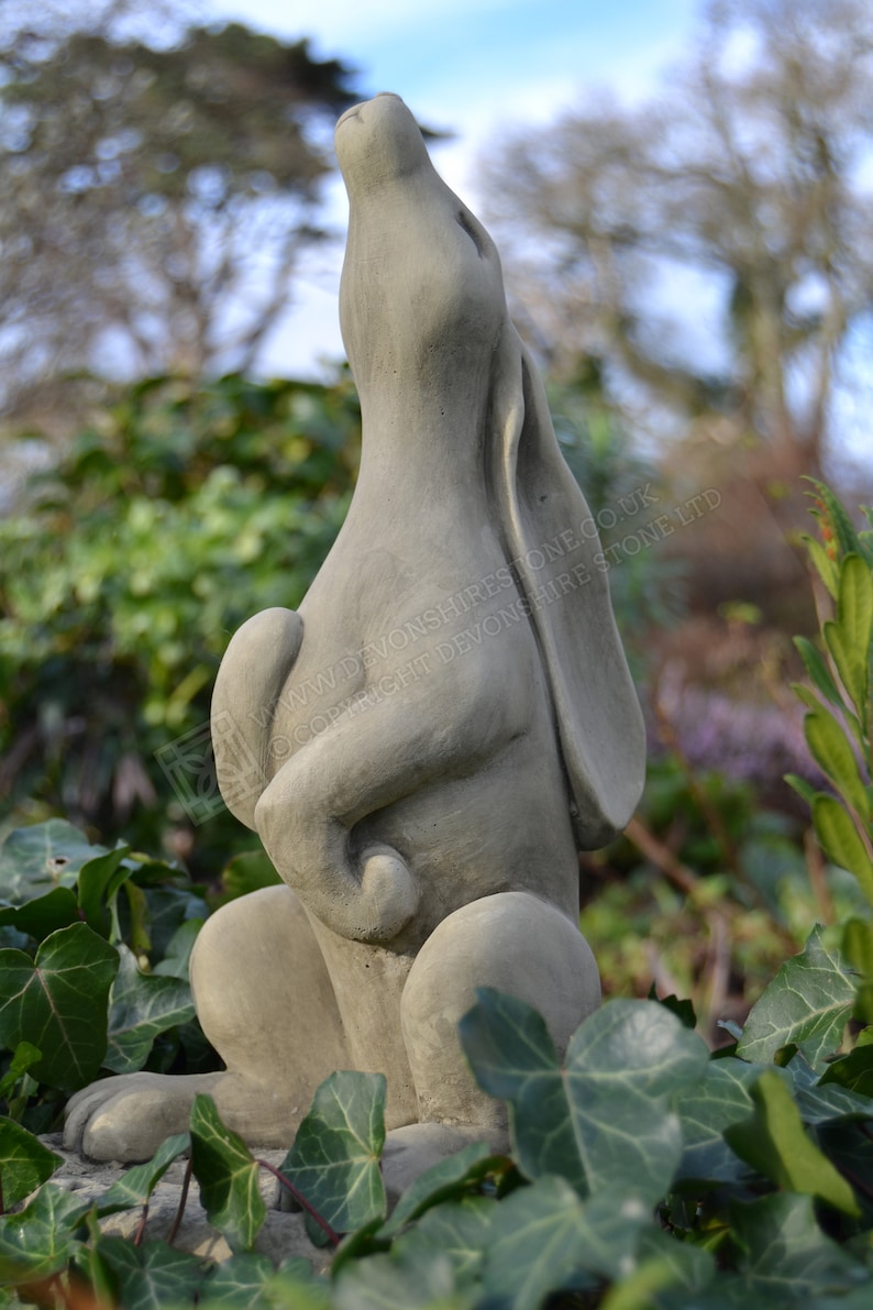 Pu&ograve; includere: Una scultura di pietra grigia di una lepre con la testa inclinata verso l'alto, che guarda verso il cielo. La scultura &egrave; seduta su un letto di foglie verdi.