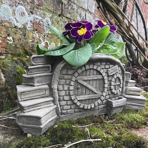 Puede incluir: Un macetero de hormigón gris en forma de casa en miniatura con una puerta y una fachada de ladrillo. El macetero está decorado con libros apilados y tiene una pequeña maceta en el lateral. El macetero está lleno de flores moradas y amarillas.