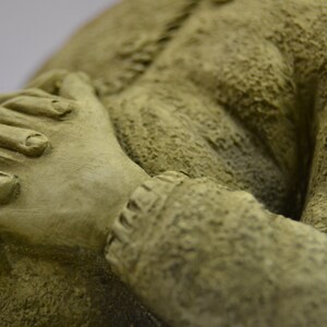 Puede incluir: Primer plano de una mano esculpida de textura verde, con los dedos entrelazados. La mano forma parte de una escultura m&aacute;s grande, que posiblemente representa a una persona en oraci&oacute;n.