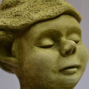 Puede incluir: Primer plano de una escultura de piedra verde de un rostro humano con los ojos cerrados. La escultura tiene una textura detallada y una nariz ligeramente hacia arriba.