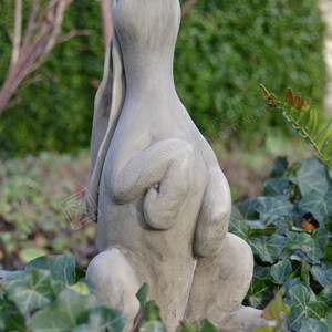 Pu&ograve; includere: Una scultura in cemento grigio di un coniglio seduto con le orecchie dritte e la testa inclinata all'indietro. La scultura &egrave; circondata da vegetazione verde.