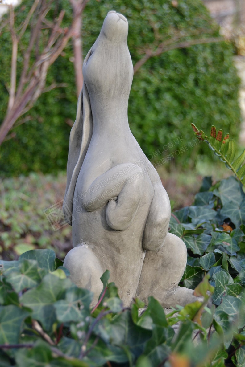 Pu&ograve; includere: Una scultura in cemento grigio di un coniglio seduto con le orecchie dritte e la testa inclinata all'indietro. La scultura &egrave; circondata da vegetazione verde.