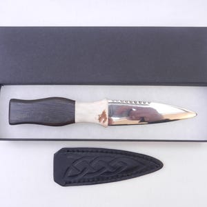 May include: A black and white handled knife with a silver blade and a serrated edge. The knife is in a black box with a white interior. The knife also comes with a black leather sheath with a Celtic knot design.