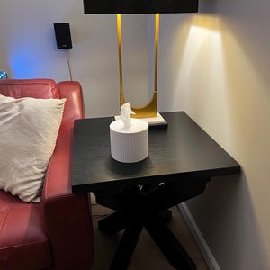 K&ouml;nnte beinhalten: Ein schwarzer quadratischer Beistelltisch mit X-f&ouml;rmigem Fu&szlig;. Eine wei&szlig;e Taschentuchbox mit vertikalen Linien steht oben. Eine goldene und schwarze Lampe befindet sich hinter der Taschentuchbox.