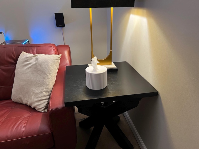 K&ouml;nnte beinhalten: Ein schwarzer quadratischer Beistelltisch mit X-f&ouml;rmigem Fu&szlig;. Eine wei&szlig;e Taschentuchbox mit vertikalen Linien steht oben. Eine goldene und schwarze Lampe befindet sich hinter der Taschentuchbox.