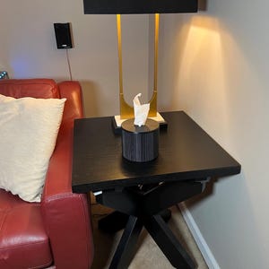 K&ouml;nnte beinhalten: Ein schwarzer quadratischer Beistelltisch mit einem Kreuzfu&szlig;. Eine schwarze Taschentuchbox mit vertikalen Linien steht oben. Eine goldene und schwarze Lampe mit schwarzem Lampenschirm steht auf dem Tisch. Ein rotes Ledersofa und ein cremefarbenes Kissen sind im Hintergrund.