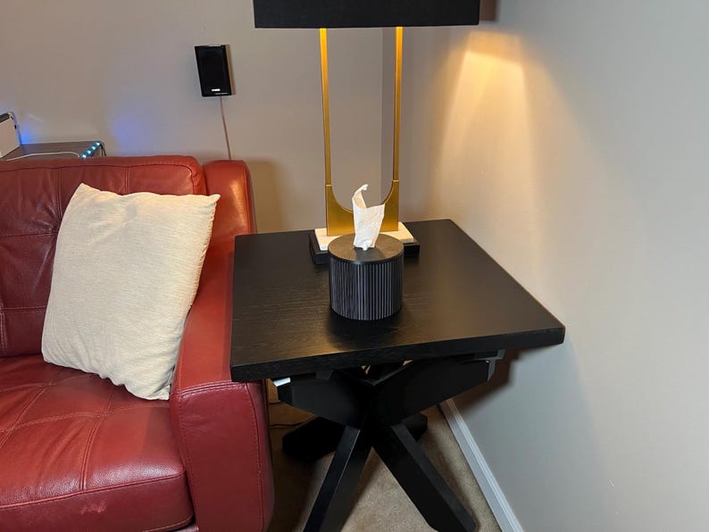 K&ouml;nnte beinhalten: Ein schwarzer quadratischer Beistelltisch mit einem Kreuzfu&szlig;. Eine schwarze Taschentuchbox mit vertikalen Linien steht oben. Eine goldene und schwarze Lampe mit schwarzem Lampenschirm steht auf dem Tisch. Ein rotes Ledersofa und ein cremefarbenes Kissen sind im Hintergrund.