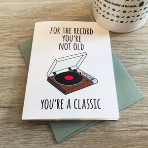 May include: A white card with a black and brown illustration of a record player. The text on the card reads "For the record you're not old you're a classic".