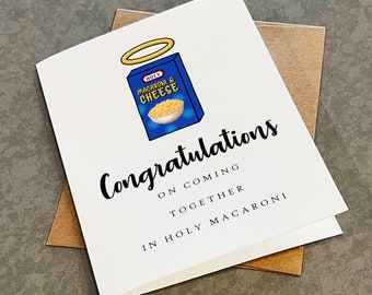 May include: A white card with a blue box of macaroni and cheese with a halo above it. The text on the card reads "Congratulations on coming together in Holy Macaroni".