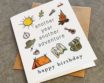 May include: A white card with a black and white illustration of camping gear, including a tent, a compass, a map, a campfire, a guitar, a flashlight, a mug, a hat, and a backpack. The text on the card reads "another year another adventure happy birthday".