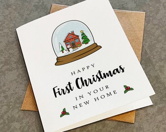 May include: A white greeting card featuring a snow globe illustration with a house, snowman, and tree inside. The text reads: HAPPY First Christmas IN YOUR NEW HOME. Two small holly sprigs with red berries are printed near the bottom corners.