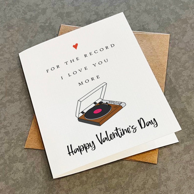 May include: A white card with a red heart and a black and brown illustration of a record player. The text on the card reads "For the record I love you more Happy Valentine's Day."