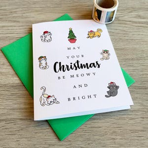 May include: A white greeting card with a green envelope featuring a central Christmas tree illustration surrounded by six cartoon cats wearing Santa hats. The text reads: MAY YOUR Christmas BE MEOWY AND BRIGHT. The card has a textured paper finish.