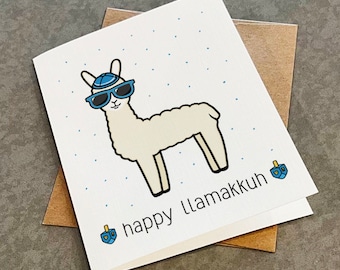 May include: A white greeting card featuring a cartoon llama wearing a blue yarmulke and blue sunglasses. The card has blue dots in the background and the text "happy LLAMAKKUH" with two blue dreidels on either side of the text.