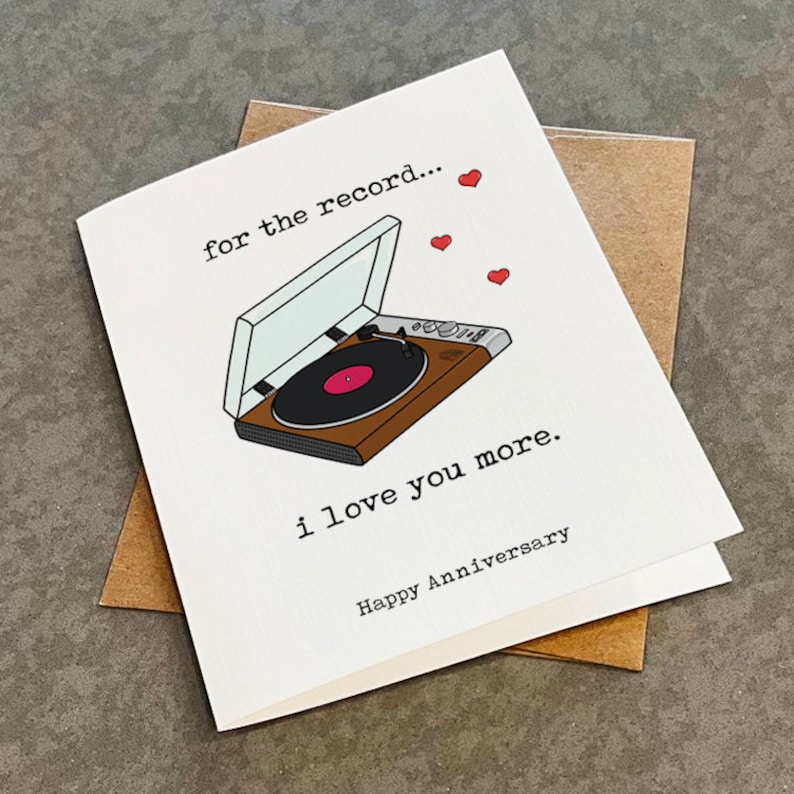 Puede incluir: Una tarjeta blanca con una ilustraci&oacute;n en negro y marr&oacute;n de un tocadiscos con el texto "for the record..." e "i love you more." La tarjeta tambi&eacute;n tiene corazones rojos y el texto "Happy Anniversary".