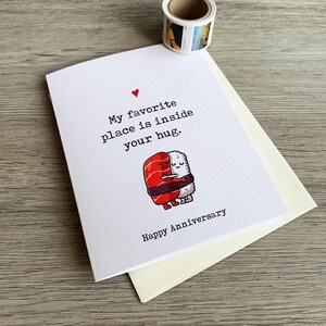 May include: A white card with a red heart and the text "My favourite place is inside your hug. Happy Anniversary."  A cartoon image of two sushi rolls hugging each other.