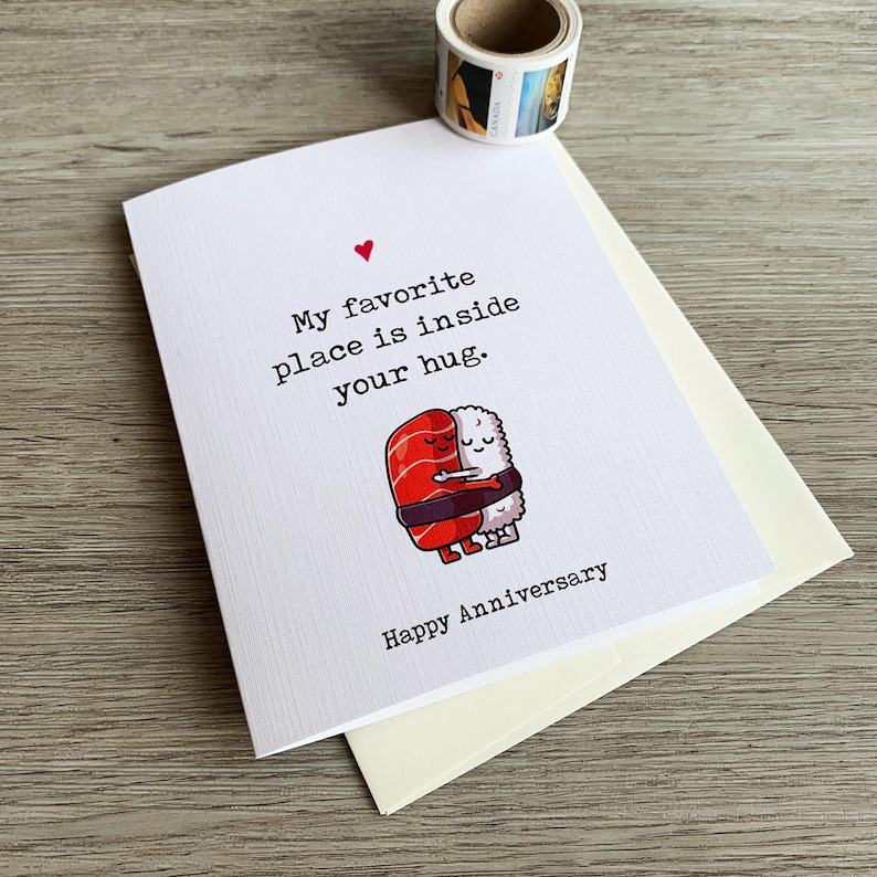 May include: A white card with a red heart and the text "My favourite place is inside your hug. Happy Anniversary."  A cartoon image of two sushi rolls hugging each other.