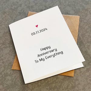 May include: A white anniversary card with a red heart and the date 03.17.2024 printed at the top. The message "Happy Anniversary To My Everything" is printed below. The card is on top of a brown envelope.