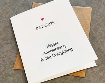 May include: A white greeting card with a small red heart icon above the date 03.17.2024, followed by the black printed text Happy Anniversary To My Everything, resting on top of a brown paper envelope.